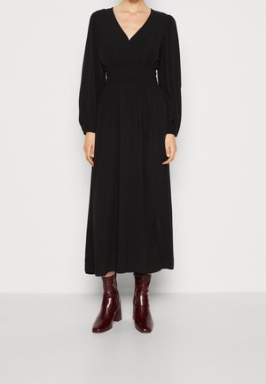 Kobieta ubrana w długą czarną sukienkę z długim rękawem, dekoltem w kształcie litery V i marszczonym pasem, zestawioną z błyszczącymi ciemnoczerwonymi botkami do kostki.