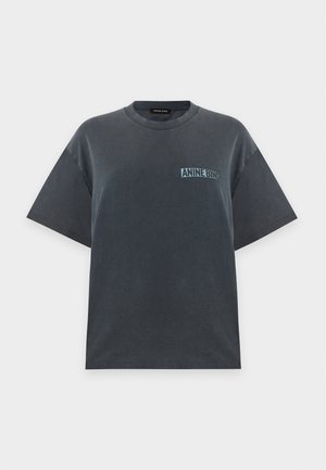 Kobieta ubrana w ciemny oversize t-shirt, jasne szorty z denimu, czarne wąskie baleriny i trzyma czarną kopertówkę na jednolitym jasnym tle.