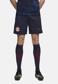 Pantalones cortos de fútbol azul marino con el logo de Red Bull Salzburg, combinados con medias azul marino hasta la rodilla que presentan el texto rojo "SOIZBURG" y el logo de Puma. Botas negras con detalles de diseño.