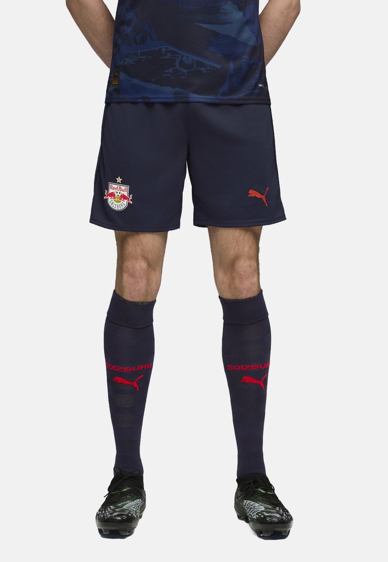 Temno modri nogometni šortai z logotipom Red Bull Salzburg, v kombinaciji z dolgimi temno modrimi nogavicami, ki nosijo rdeče besedilo "SOIZBURG" in logotip Puma. Črne kopačke z oblikovalskimi poudarki.