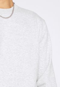Sudadera gris con cuello redondo, con un tejido suave y texturizado y un collar de cadena plateada visible en la zona del cuello.