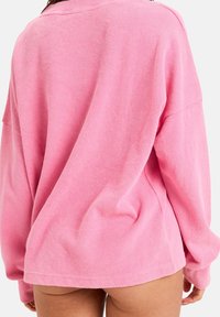Przesadnie duża różowa bluza z teksturowanego materiału, z długimi rękawami i luźnym krojem, z zaokrąglonym dekoltem i bocznymi rozcięciami.