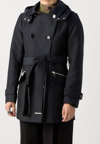 Manteau bleu marine avec un design croisé, quincaillerie en argent brillant, capuche ajustable et deux poches avant zippées. Comprend une taille ceinturée.