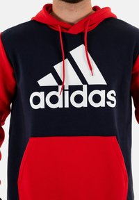 Felpa con cappuccio navy e rossa in misto cotone, caratterizzata da un grande logo Adidas bianco e una tasca a marsupio sul davanti. Tessuto texture con cordini.