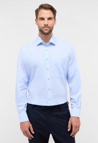 Camicia azzurro chiaro a maniche lunghe con colletto a punta, tasca frontale e dettagli con bottoni. Il tessuto sembra liscio e leggermente texturizzato.