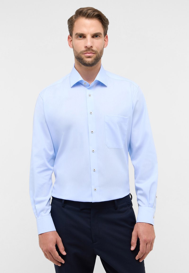 Camicia azzurro chiaro a maniche lunghe con colletto a punta, tasca frontale e dettagli con bottoni. Il tessuto sembra liscio e leggermente texturizzato.