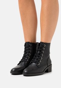 Bottes en cuir noir à la cheville avec fermeture à lacets, œillets argentés et talon bloc. Le design présente une texture lisse et un détail zippé.