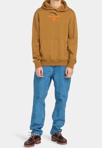 Brown hoodie with front kangaroo pocket and orange logo, paired with light blue denim jeans and dark brown shoes with lace details.