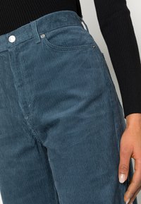 Velourkové kalhoty v temně modré barvě; mají vertikální ribkovou strukturu a design s pěti kapsami s kovovými detaily.