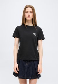 Γυναίκα με ίσια ανοιχτό καστανά μαλλιά, που φοράει μαύρο t-shirt Calvin Klein και σκουρόχρωμη ραμμένη τζιν φούστα, μπροστά σε απλό φόντο.