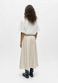 Femme aux cheveux longs et bouclés portant un t-shirt blanc surdimensionné, une jupe midi beige, et des bottes noires montant jusqu'au milieu du mollet, vue de dos.