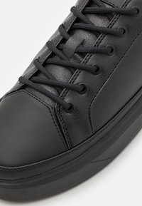 Musta synteettinen nahkainen lenkkari, jossa on teksturoitu päälliosa, pyöreä kärki, kuusi nauhanreikää ja tasaiset mustat nauhat. Varustettu paksulla kumipohjalla.