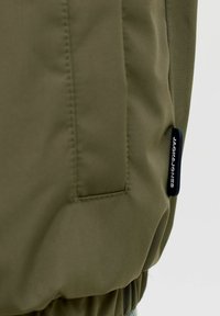 Κοντινό πλάνο στην τσέπη ενός ελιάς πράσινου μπουφάν με κατακόρυφη ραφή και μικρή μαύρη υφασμάτινη ετικέτα με την επιγραφή "JACK & JONES" στο πλάι.