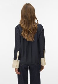 Camisa azul marino oscuro de manga larga, con puños beige claro, tela texturizada y detalles de botones. Mostrada desde atrás, destacando el cuello y la longitud.