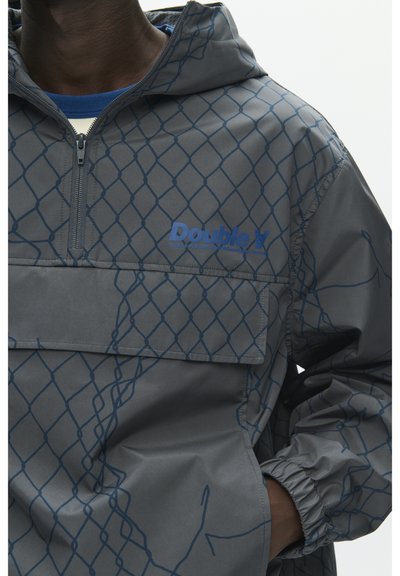 Veste à capuche grise avec motif de chaîne bleue, fermeture éclair partielle, poche avant à rabat et texte bleu "Doublet" sur la poitrine, portée par une personne avec la main dans la poche.