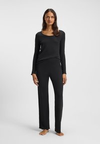 Γυναίκα όρθια, με γυμνά πόδια, φοράει μαύρο μακρυμάνικο τοπ και μαύρο χαλαρό παντελόνι, σε απλό ανοιχτό φόντο.