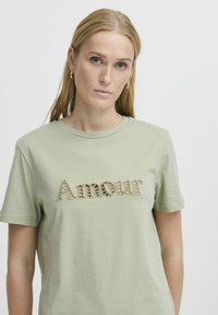 Světle zelené bavlněné tričko s texturovaným, vyšívaným designem "Amour" v béžové barvě. Krátké rukávy a kulatý výstřih.