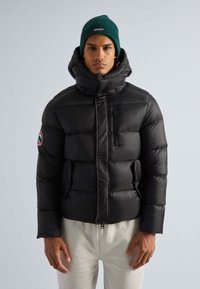 Schwarze Puffjacke mit hohem Kragen, Reißverschlusstaschen und Logo-Patch. Getragen mit einer grünen Strickmütze. Glattes, leichtes Material, gestepptes Design.