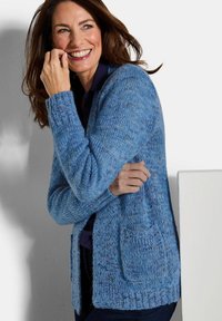 Lächelnde Frau mit langen braunen Haaren, die eine blaue Strickjacke über einem dunklen Oberteil trägt, vor einem einfarbigen hellen Hintergrund stehend.