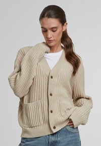 Cardigan tricotat bej cu decolteu în V, închidere cu nasturi și două buzunare frontale. Textură ribbed cu detalii decorative de cusături.