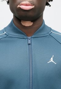 Μπλε αθλητικό μπουφάν με φερμουάρ μπροστά, με λευκές λεπτομέρειες και λογότυπο Jumpman στο στήθος. Κατασκευασμένο από λείο, ελαστικό υλικό.