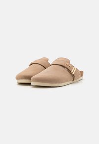 Beige semskede slip-on sko med rund tå, bred stropp over toppen, og en gullspenne detalj. myk tekstur med flat såle.