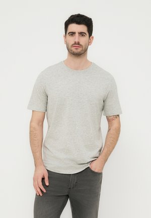 Άνδρας με σκούρα μαλλιά, φορώντας ανοιχτό γκρι κοντομάνικη μπλούζα και σκούρα γκρι τζιν, στέκεται μπροστά σε λευκό φόντο με το ένα χέρι στην τσέπη.