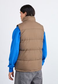 Rjava puff vest z visokim ovratnikom, prešito zasnovo in podrobnostjo na hrbtu, oblečen preko modre srajce z dolgimi rokavi in sivih hlač.