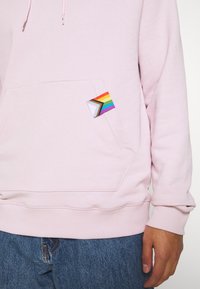 Felpa con cappuccio rosa chiaro realizzata in tessuto morbido, con tasca frontale decorata da un triangolo arcobaleno. Presenta polsini e orlo a costine.