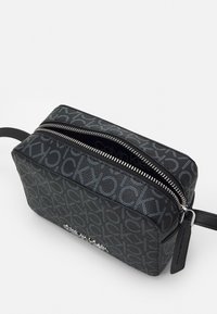 Borsa da toilette rettangolare nera in materiale sintetico strutturato. Presenta chiusura con zip color argento e motivo logo ripetuto. Tracolla nera fissata.