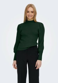 Dark green ribbed knit sweater with a high neck, long puff sleeves, and a fitted silhouette, worn paired with black trousers.