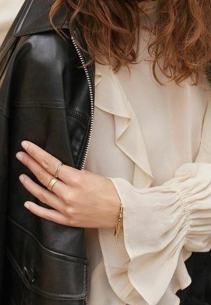 Main avec des bagues en or et un bracelet reposant sur une veste en cuir noire portée sur un chemisier crème à manches volantées et des boucles lâches.