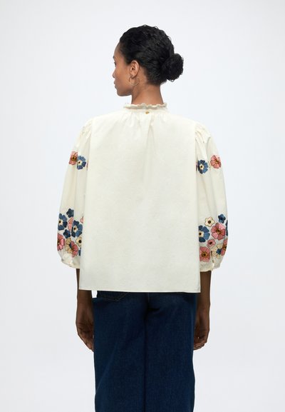 Femme aux cheveux foncés en chignon portant un chemisier blanc avec une broderie florale colorée sur les manches bouffantes et un jean bleu foncé, de dos.