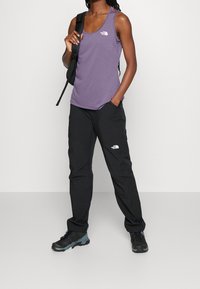 Fioletowy bezrękawnik, czarne spodnie z kieszeniami i buty trekkingowe. Top ma logo, a spodnie mają podobne logo na nogawce.