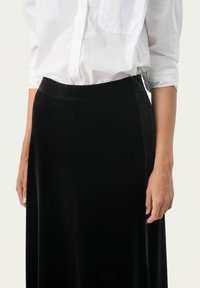 Schwarzer Samtrock mit glatter Textur, hoher Taille und bodenlangem Design. Kombiniert mit einem weißen Hemd mit Knöpfen.