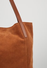 Sac fourre-tout en daim marron avec coutures surpiquées et anse en cuir lisse sur un fond gris clair uni.