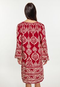 Rochie cu mânecă lungă, până la genunchi, în nuanțe de roșu și crem. Prezintă modele florale și geometrice complexe, cu manșete elastice și un guler plisat.