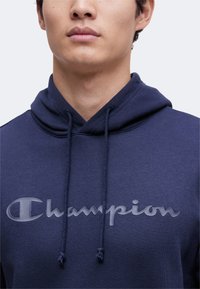 Άνδρας που φοράει μπλε σκούρο φούτερ Champion με κορδόνια και γκρίζο λογότυπο στο κέντρο του στήθους, εμφανίζεται από τον λαιμό έως τον άνω κορμό.