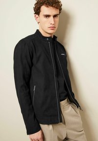 Chaqueta bomber negra con cierre frontal, cuello a presión y dos bolsillos laterales con cremallera. Presenta una textura mate elegante y un diseño ajustado.