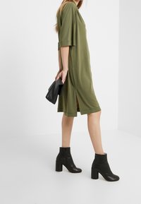 Robe vert olive jusqu'aux genoux avec des manches courtes, fentes latérales et texture lisse, associée à des bottines noires et une petite pochette noire.
