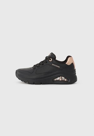 Sneaker nera Skechers con dettaglio del tallone in rosa oro, design con lacci e suola con cuscinetto d'aria visibile, mostrata su uno sfondo grigio chiaro.