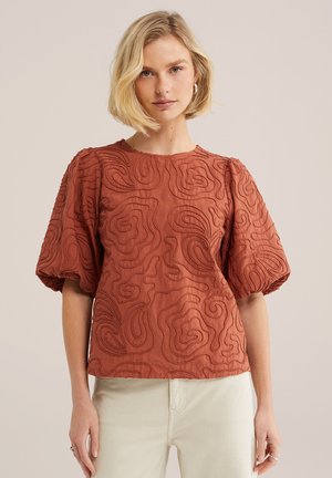 Femeie blondă purtând un top cu mâneci bufante de culoare ruginiu, decorat cu modele abstracte cusute, asortat cu pantaloni bej deschis, stând în picioare pe un fundal simplu.