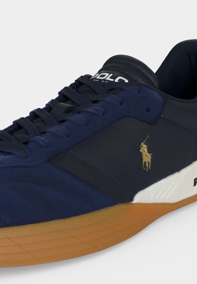 Zapatilla deportiva de cuero azul marino con suela de goma, media suela blanca, logo dorado de jugador de polo y cordones negros.