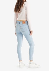 Woman wearing light blue skinny jeans, white long-sleeve cropped top, and white Levi’s sneakers, standing against a plain background.