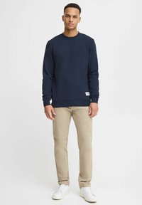 Navy blue cotton sweatshirt with a crew neck, ribbed cuffs, and hem, paired with light beige pants and white sneakers. Tag visible on the hem.