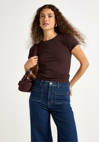 Top a maniche corte in maglia di un intenso bordeaux abbinato a jeans blu a vita alta e a gamba larga. Il modello tiene una piccola borsa a tracolla di un intenso bordeaux.
