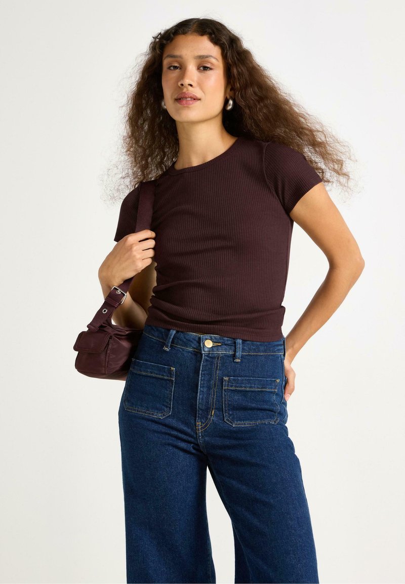Top a maniche corte in maglia di un intenso bordeaux abbinato a jeans blu a vita alta e a gamba larga. Il modello tiene una piccola borsa a tracolla di un intenso bordeaux.