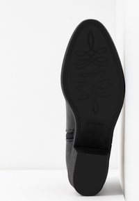 Chaussure noire avec une surface texturée, une section médiane lisse et une semelle en caoutchouc plate ornée d'un motif floral au bas. Détail de fermeture éclair sur le côté.