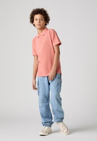 Camisa polo rosa de manga corta y cuello, combinada con pantalones cargo de color azul claro con bolsillos laterales y puños elásticos. Zapatillas blancas.
