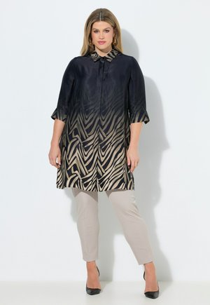 Nainen pukeutuneena mustakelta-beigeen seepra-kuvioituun tunikaan, jossa on neljänneshihat, beiget housut ja mustat korkokengät valkoista taustaa vasten.